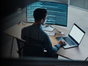 photograph from a wall camera POV of a man sitting at a desk with a laptop on the right, large desktop monitor on the left with lines of code