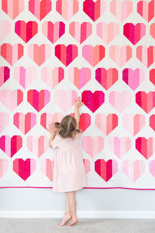 Toddler girl standing in front of heart pattern quilt hanging on wall.