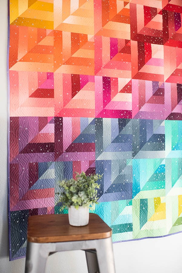 Quilt with bright modern geometric patterns and colors hanging on wall next to plant. 